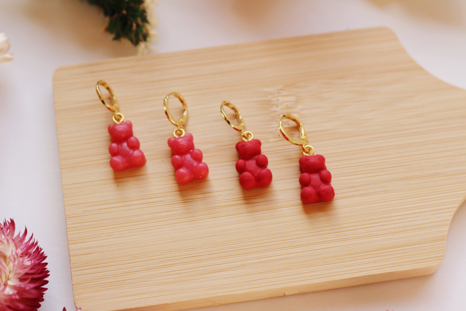 Four red gummy bear-shaped earrings with gold hooks on a wooden board.