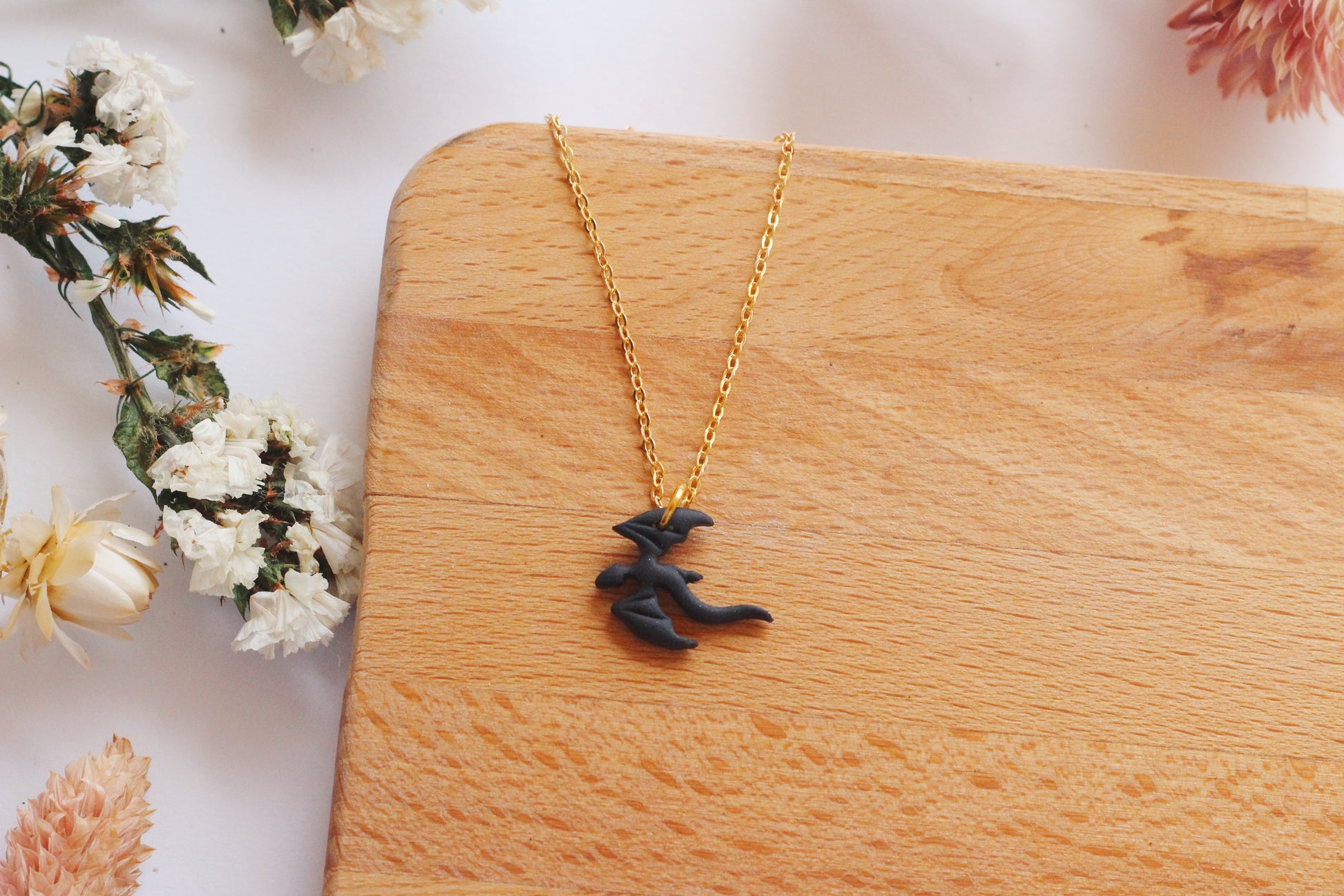 Black dragon pendant necklace on a gold chain displayed on a wooden surface with flowers in the background.