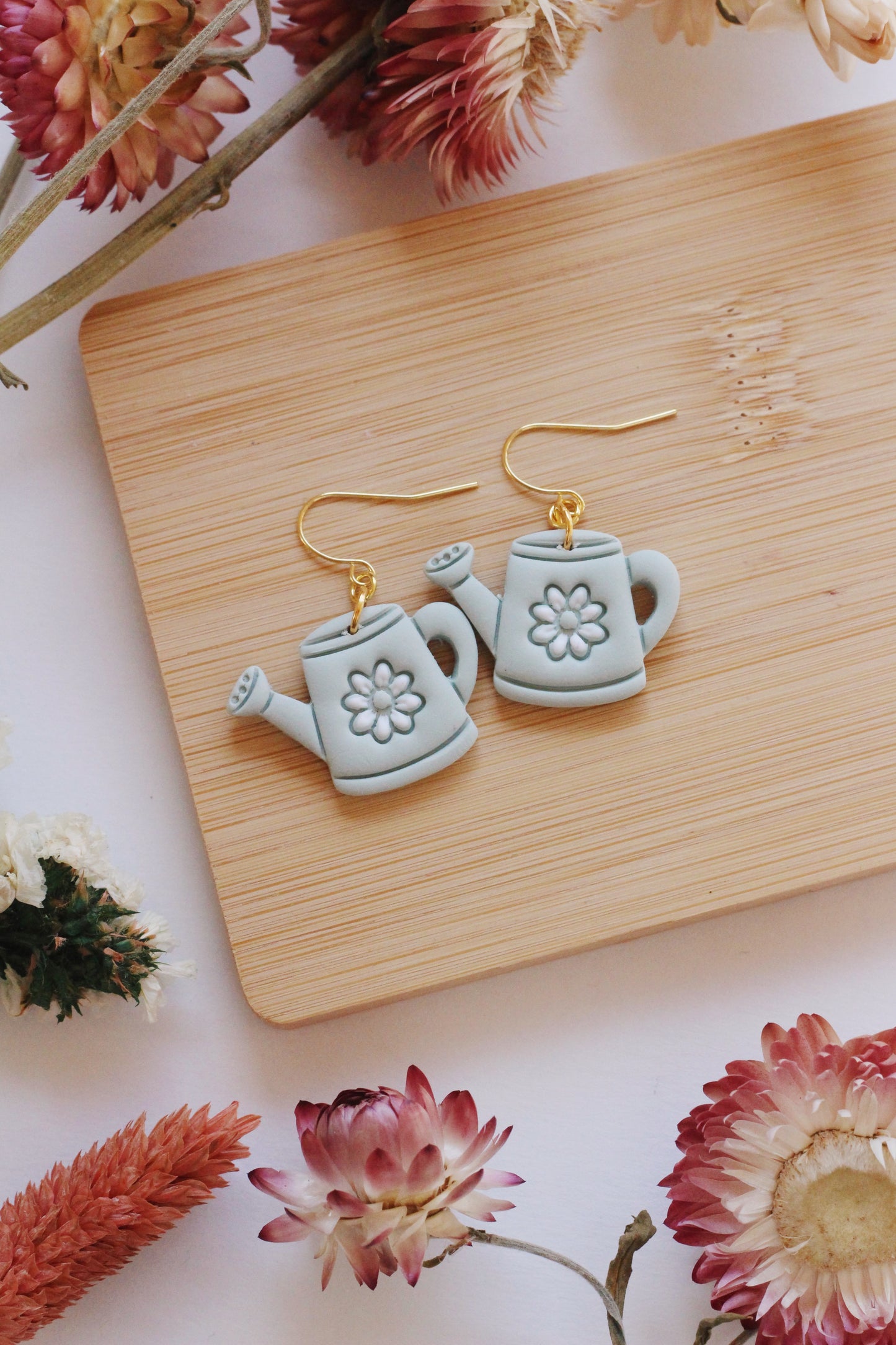 Watering can-shaped earrings on a wooden board with flowers around