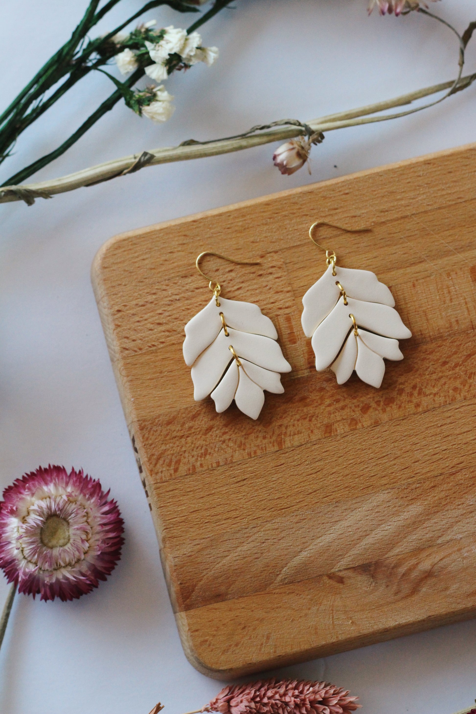 White leaf-shaped earrings on a wooden board with flowers in the background