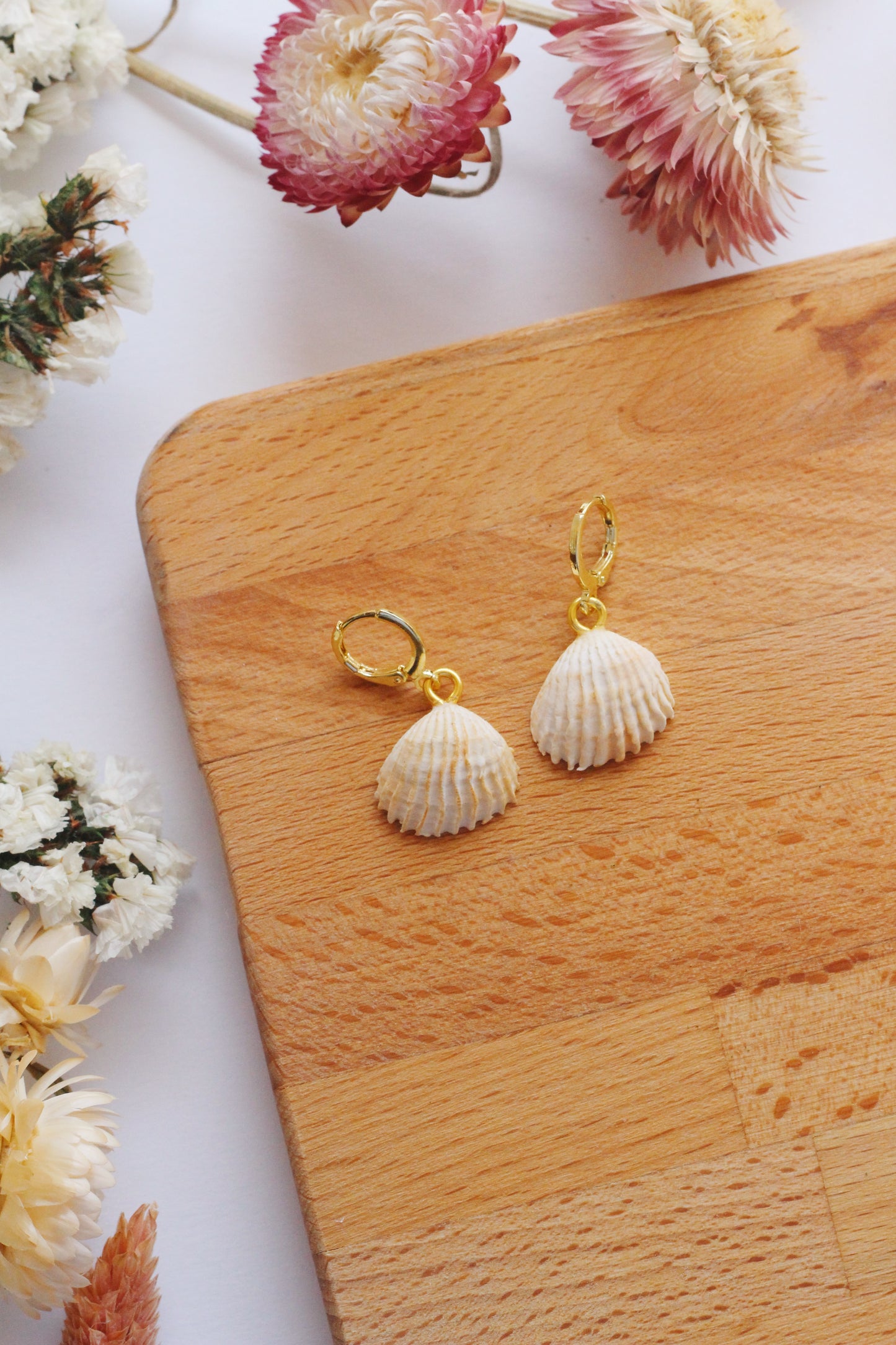 Shell-shaped earrings on a wooden board with flowers around