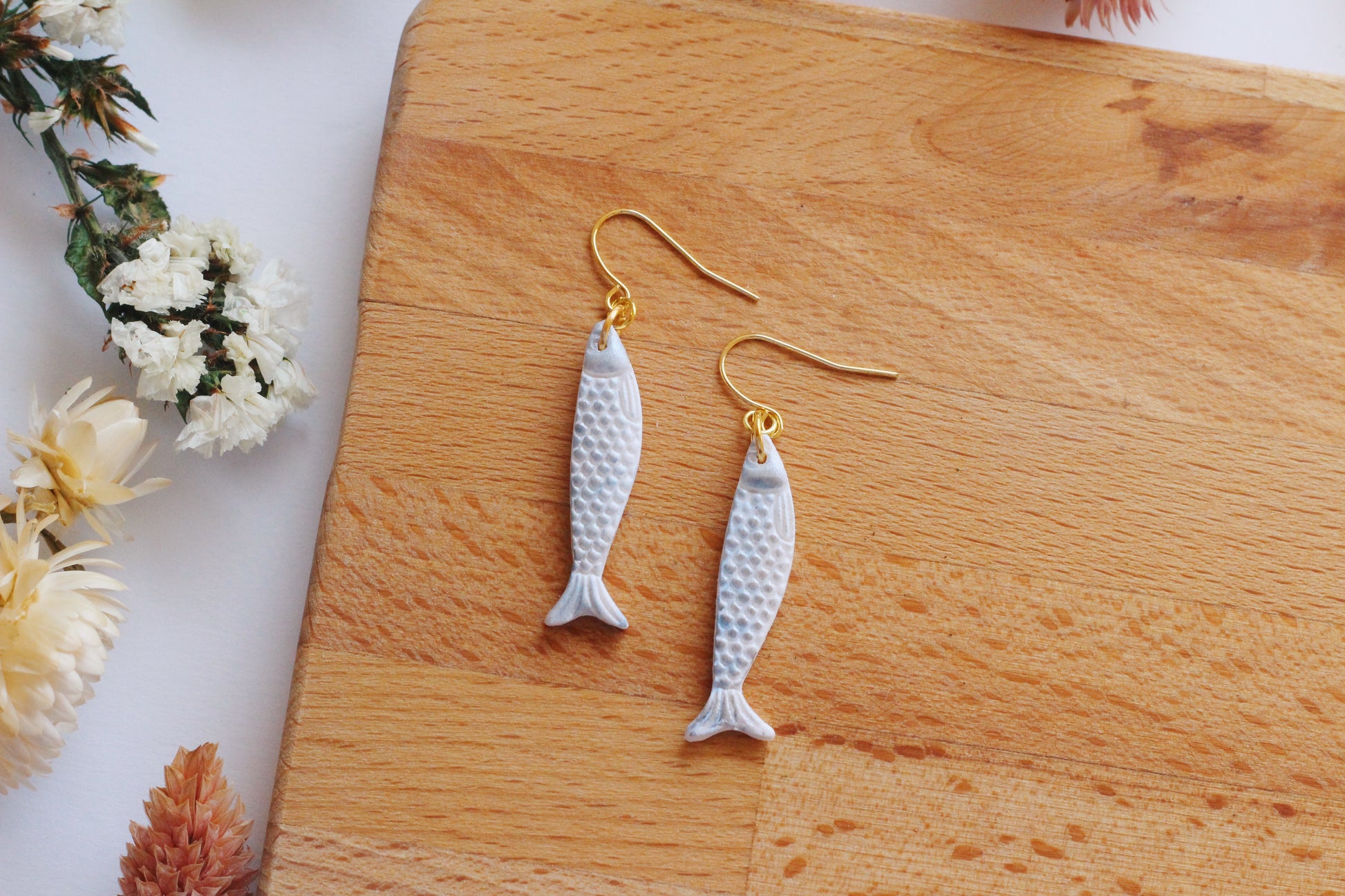 Sardine fish-shaped earrings on gold hooks displayed on a wooden board with flowers.