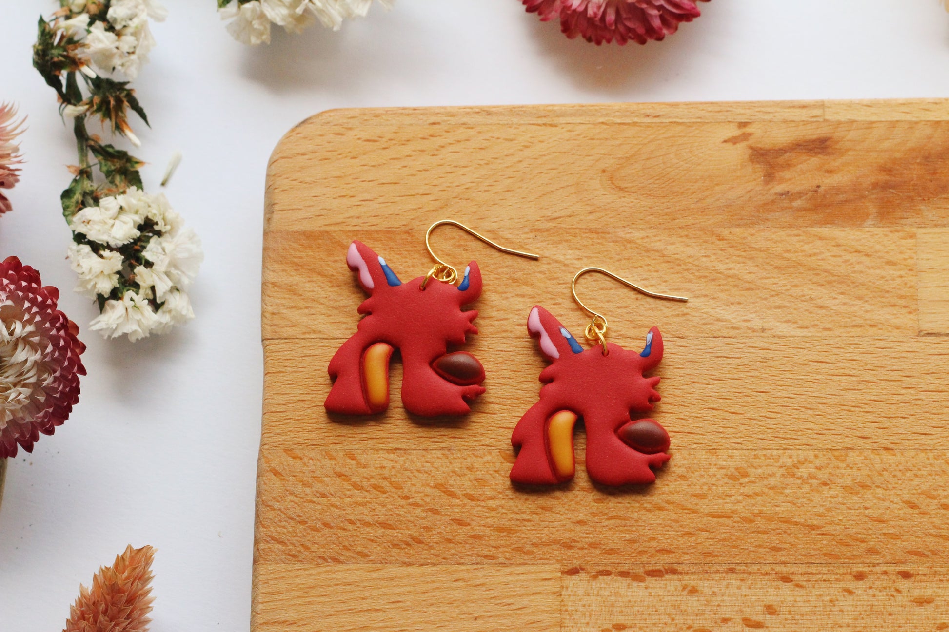 Red dragon-shaped earrings on a wooden board with flowers around