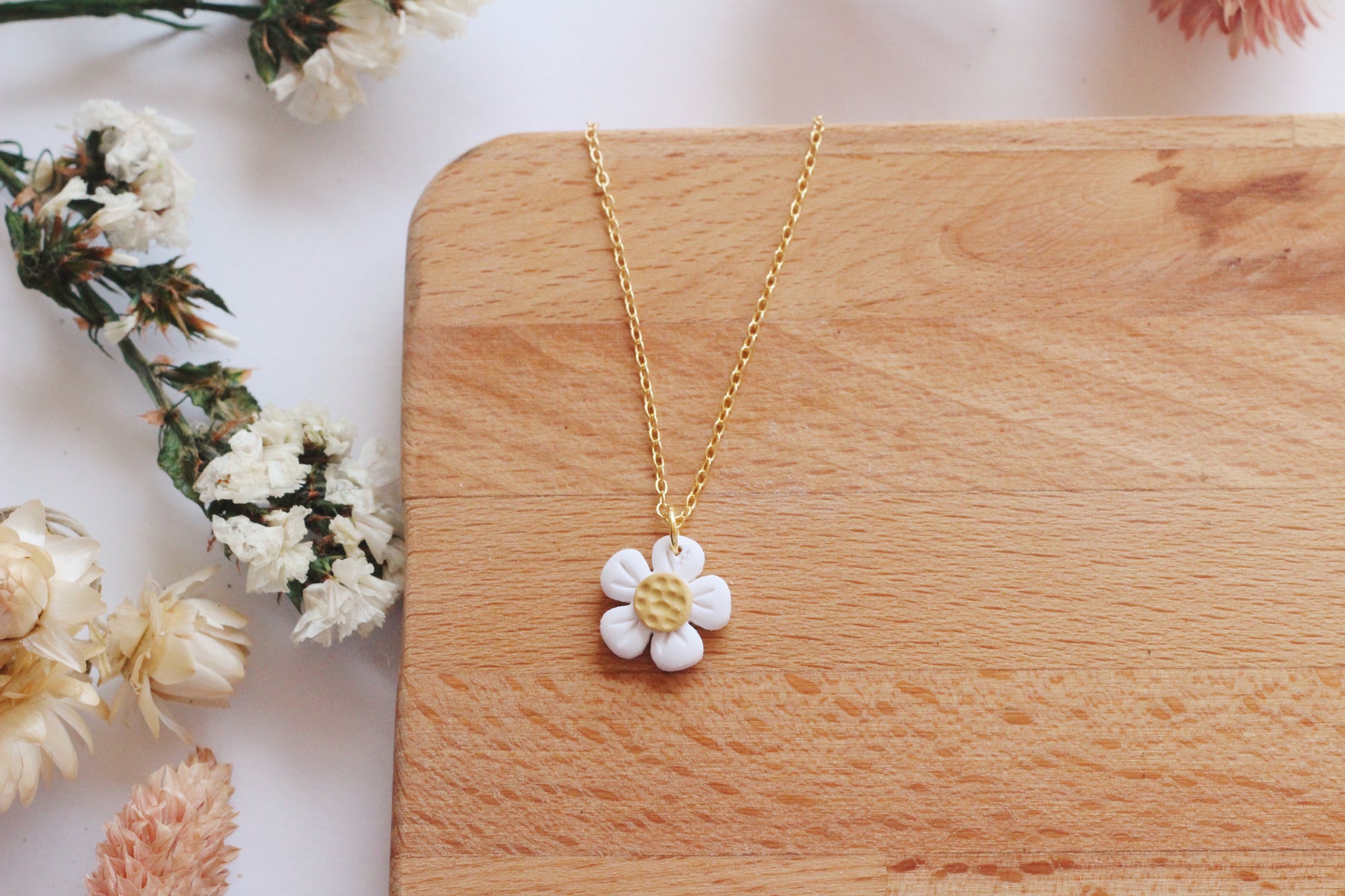 Gold necklace with a white flower pendant on a wooden surface with flowers.