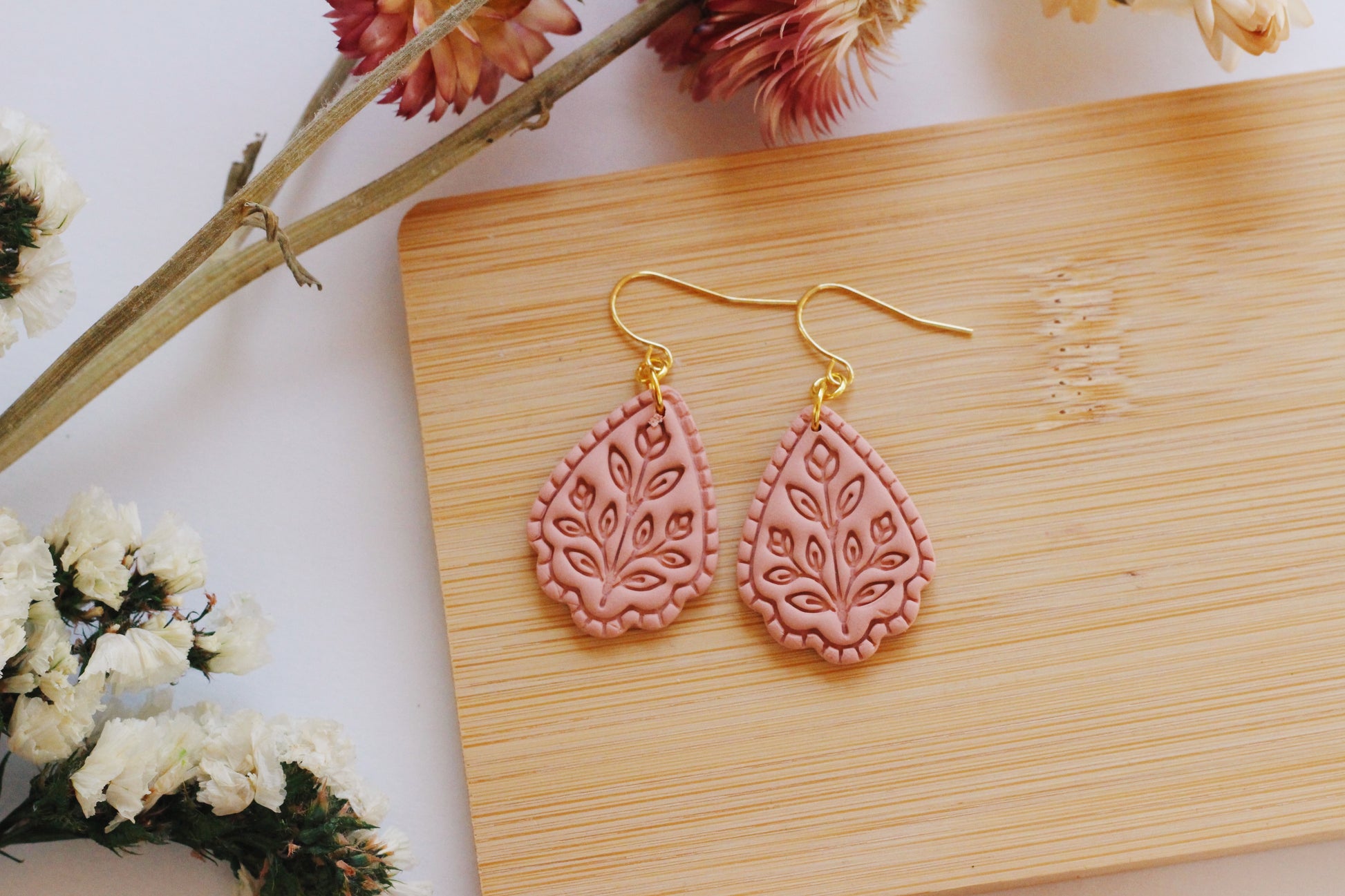 Pink teardrop earrings with gold hooks on a wooden board with flowers in the background