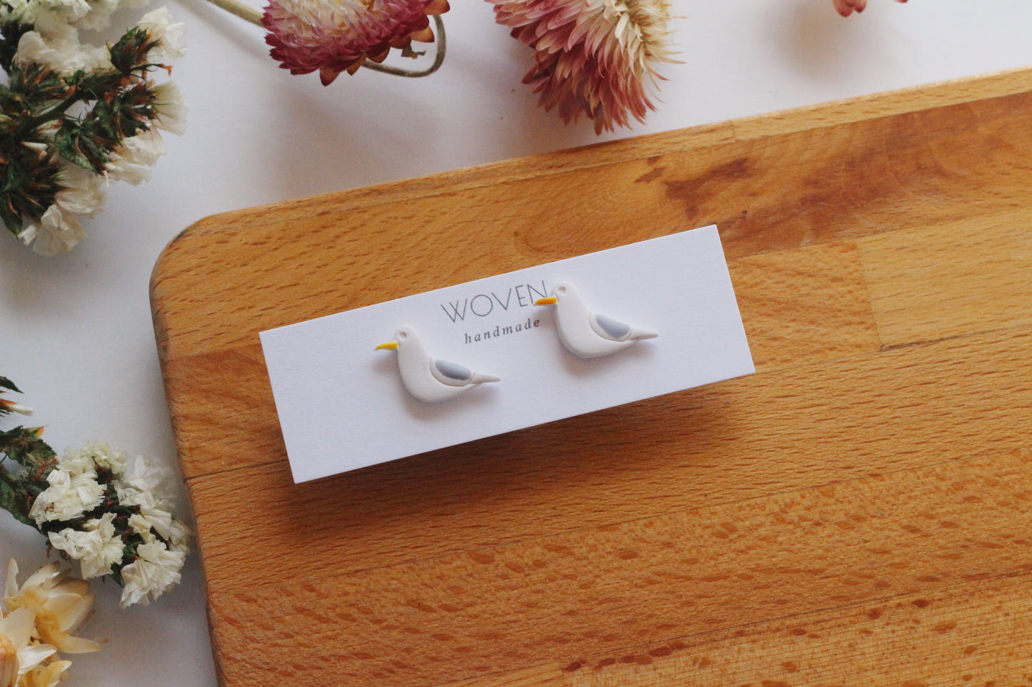 Pair of bird-shaped earrings on a 'Woven Handmade' card with dried flowers in the background.