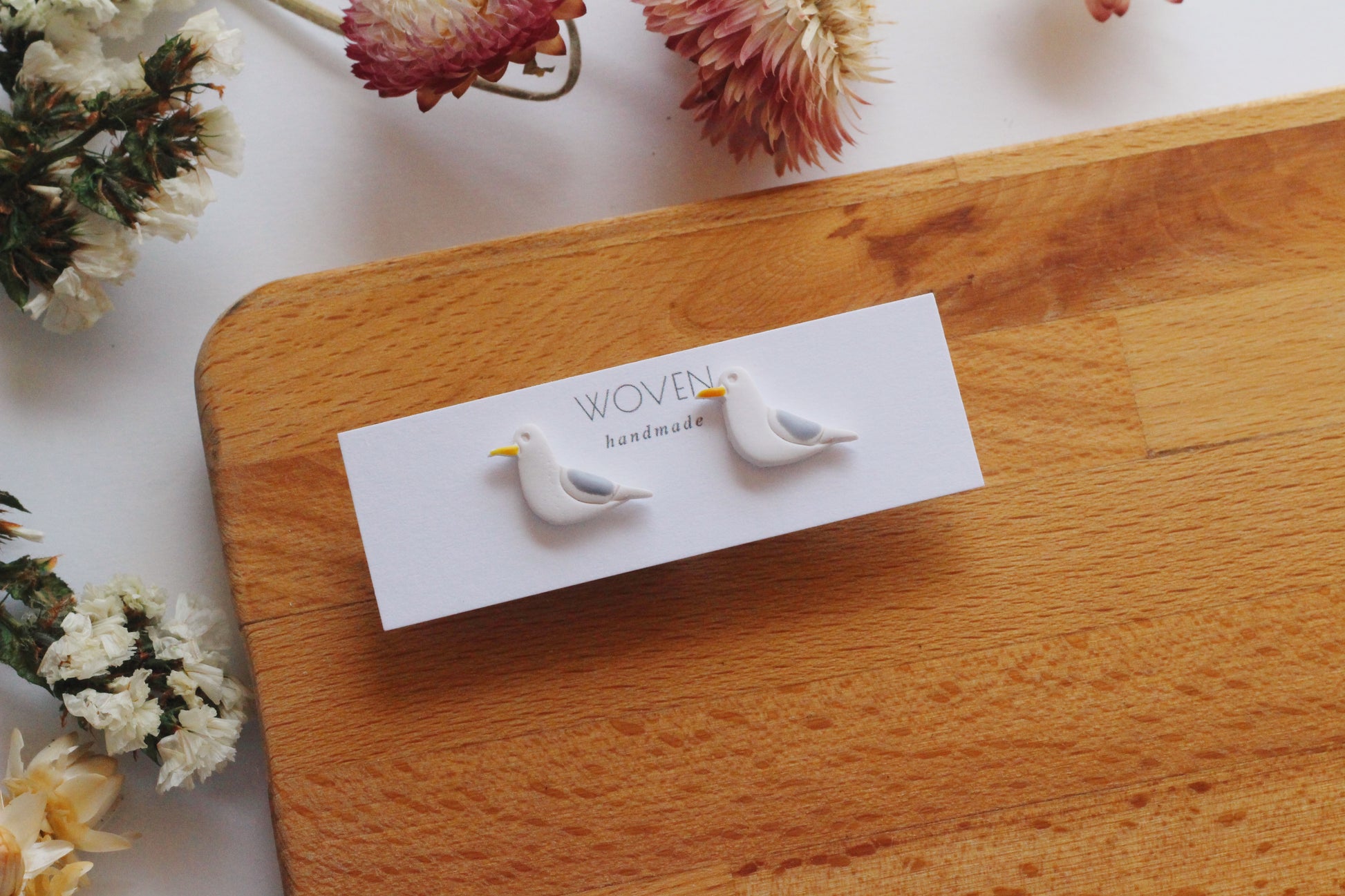 Pair of bird-shaped earrings on a 'Woven Handmade' card with dried flowers in the background.