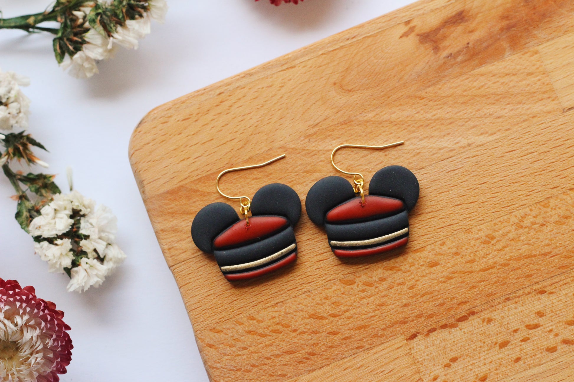 Pair of earrings mouse ear shaped like Mickey Mouse ears on a wooden surface with flowers around.