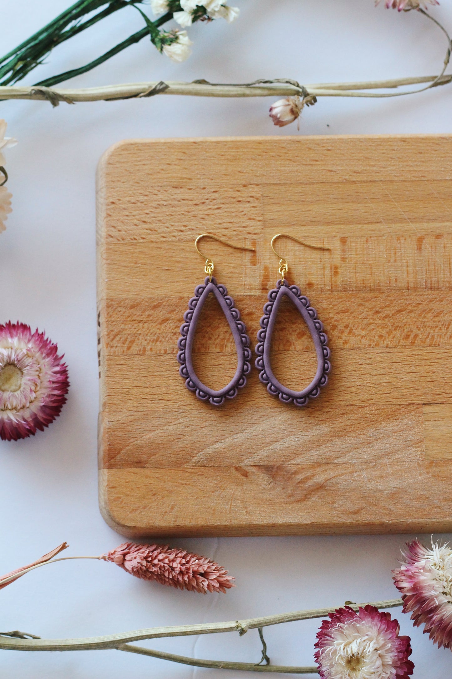Purple teardrop earrings on a wooden board with flowers around
