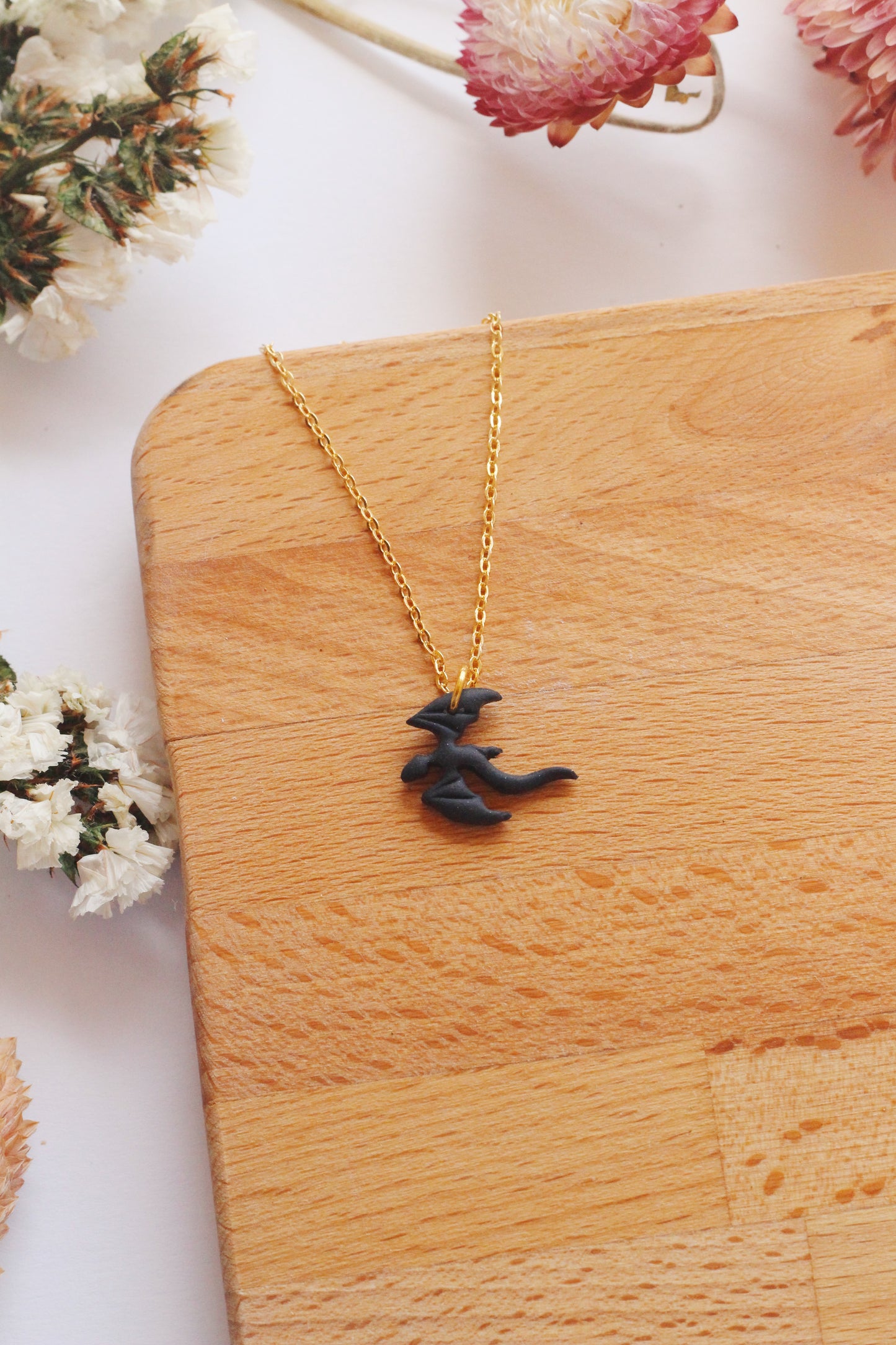 Black dragon pendant necklace on a gold chain, placed on a wooden surface with flowers in the background.