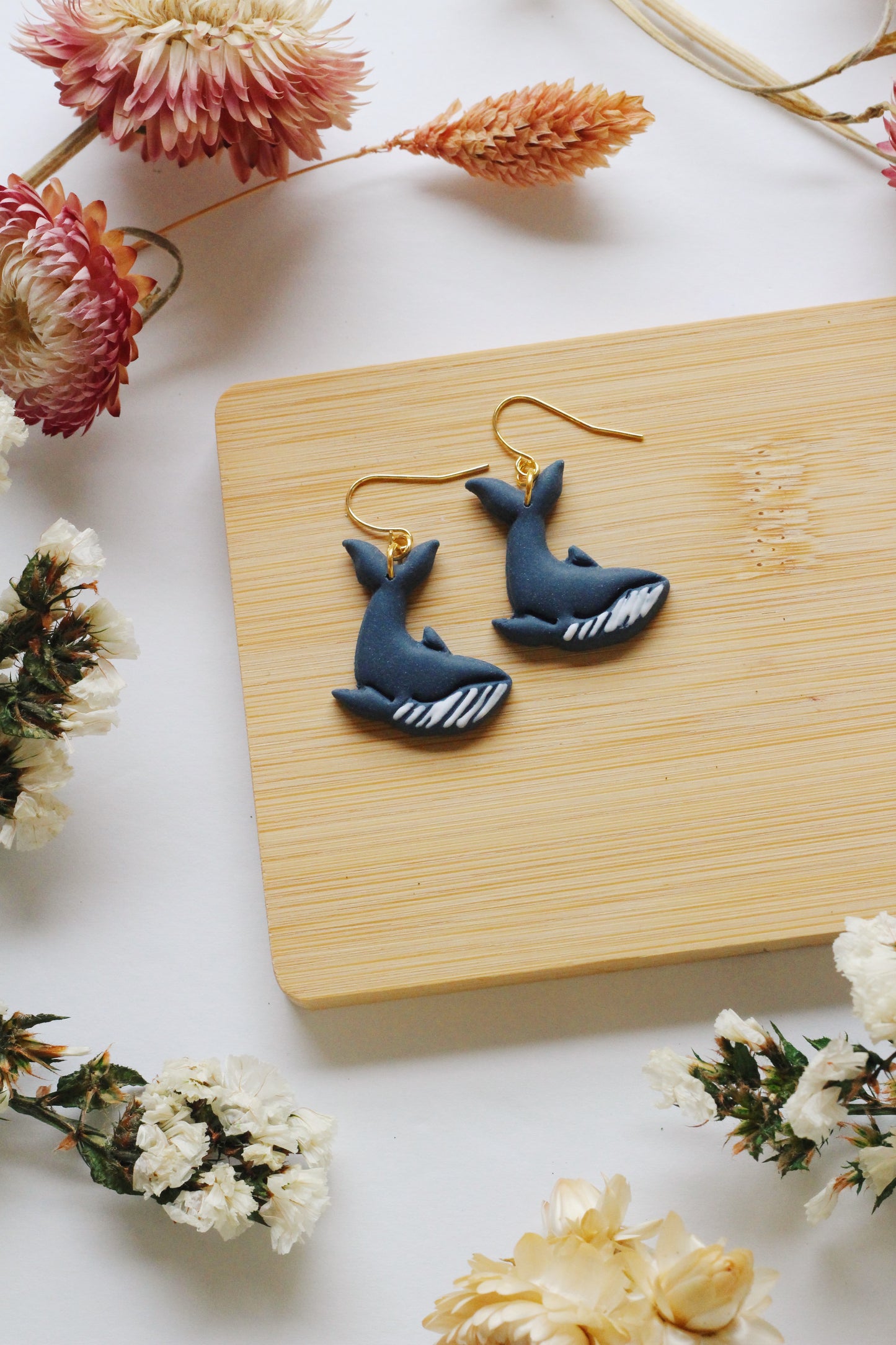 Blue whale-shaped earrings on a wooden board with flowers around
