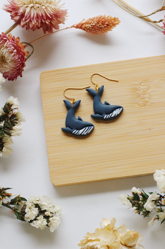 Blue whale-shaped earrings on a wooden board with flowers around
