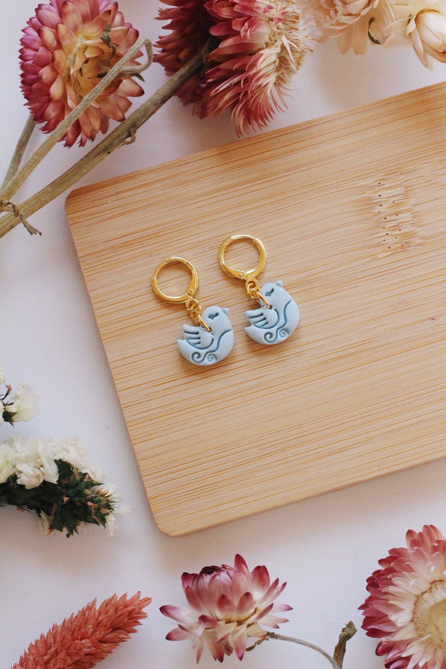 Pair of bird-shaped earrings on a wooden board with flowers around