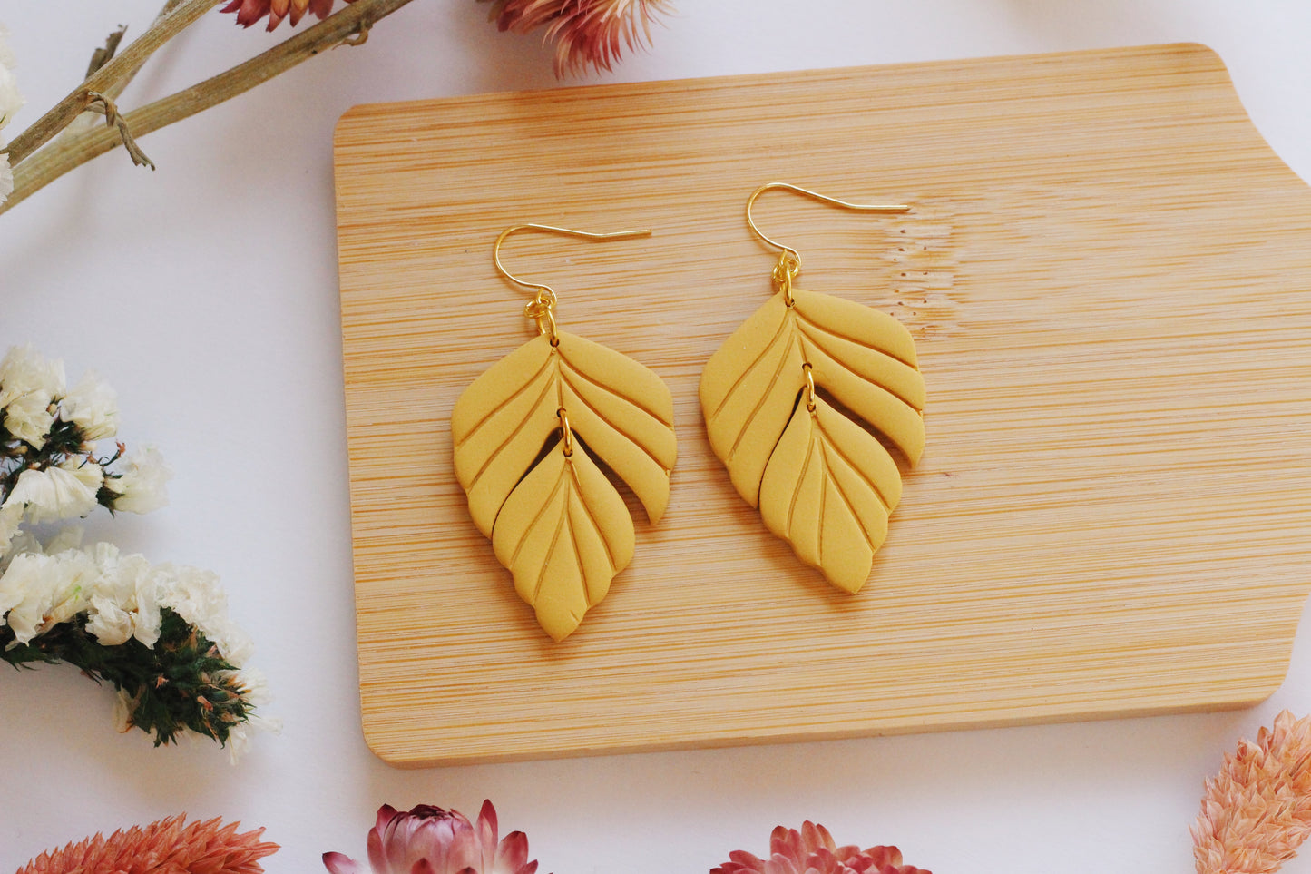 Gold leaf-shaped earrings on a wooden board with flowers around