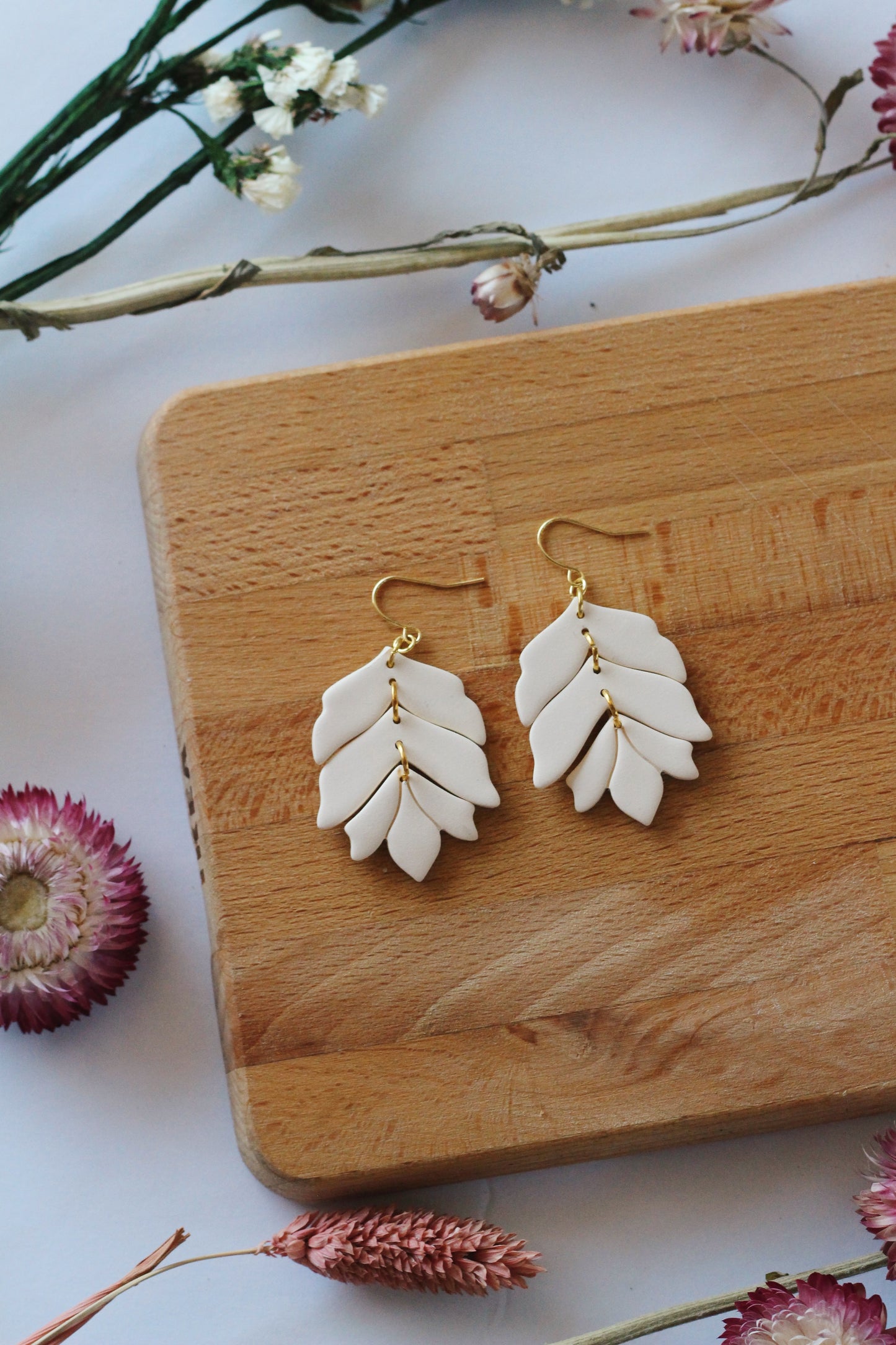 White leaf-shaped earrings on a wooden board with flowers around