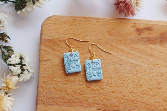 Pair of fish earrings on a wooden board with flowers in the background