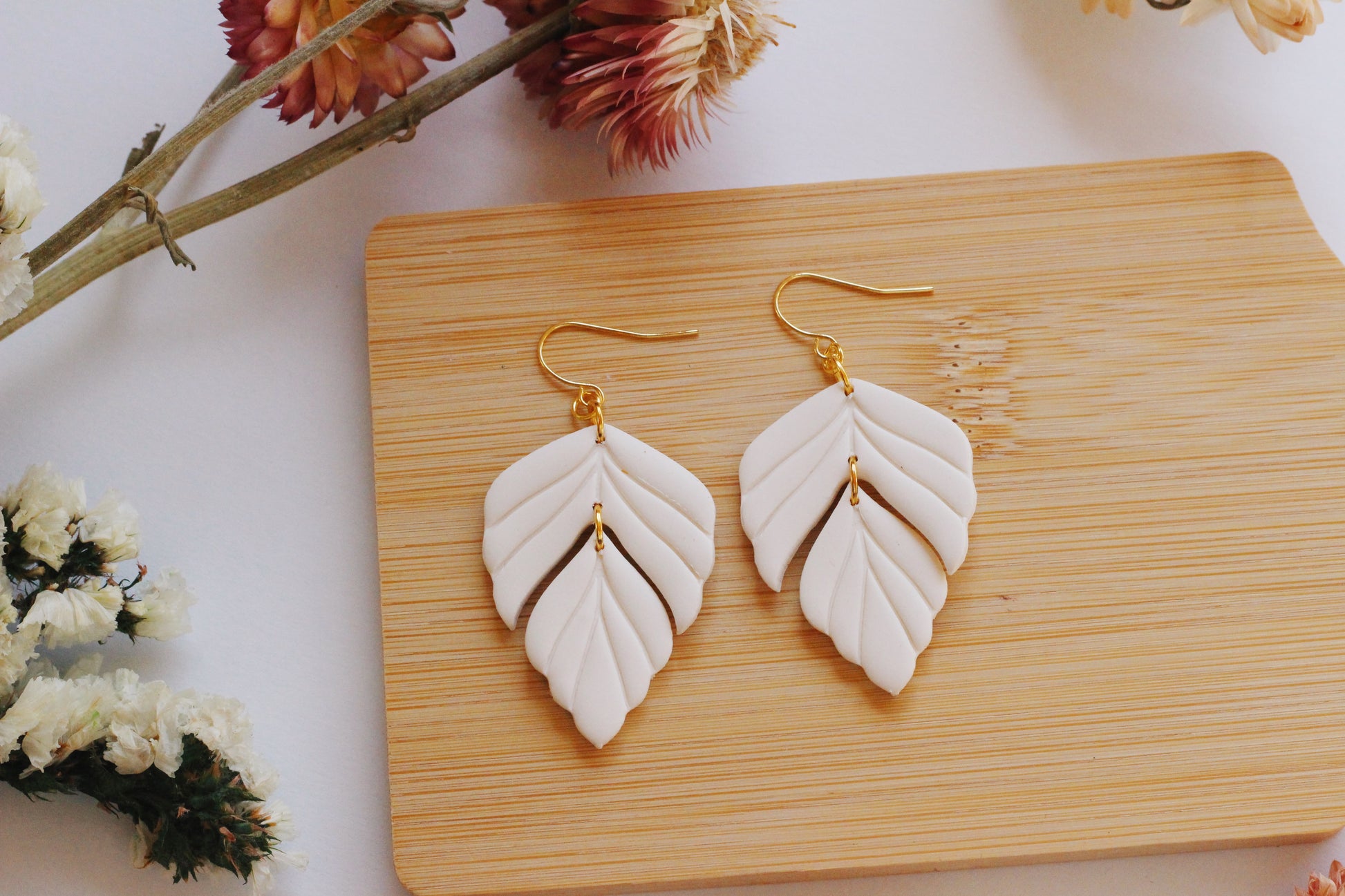 White leaf-shaped earrings on a wooden board with flowers in the background