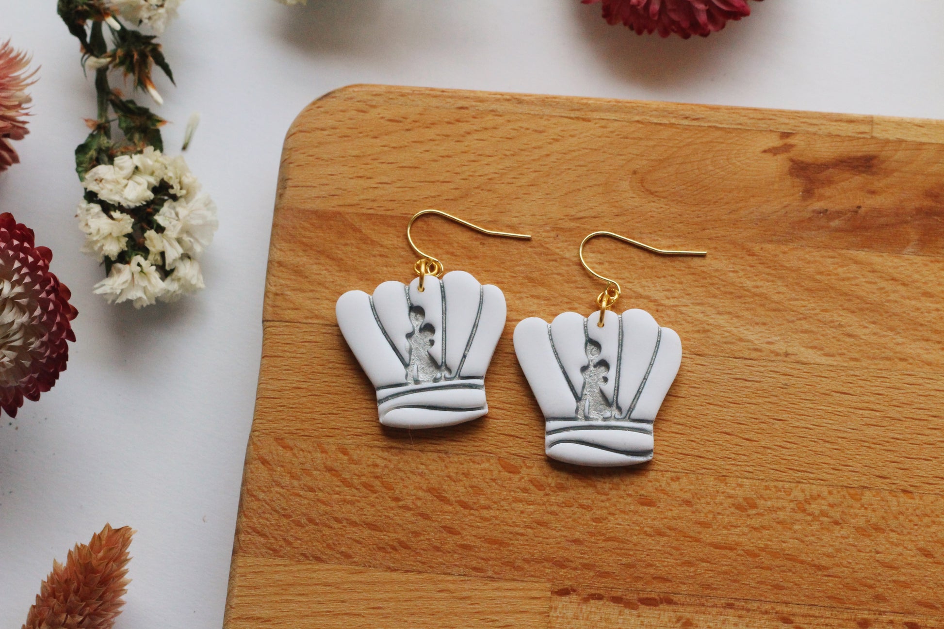 White chef earrings on gold hooks displayed on a wooden board with flowers in the background.