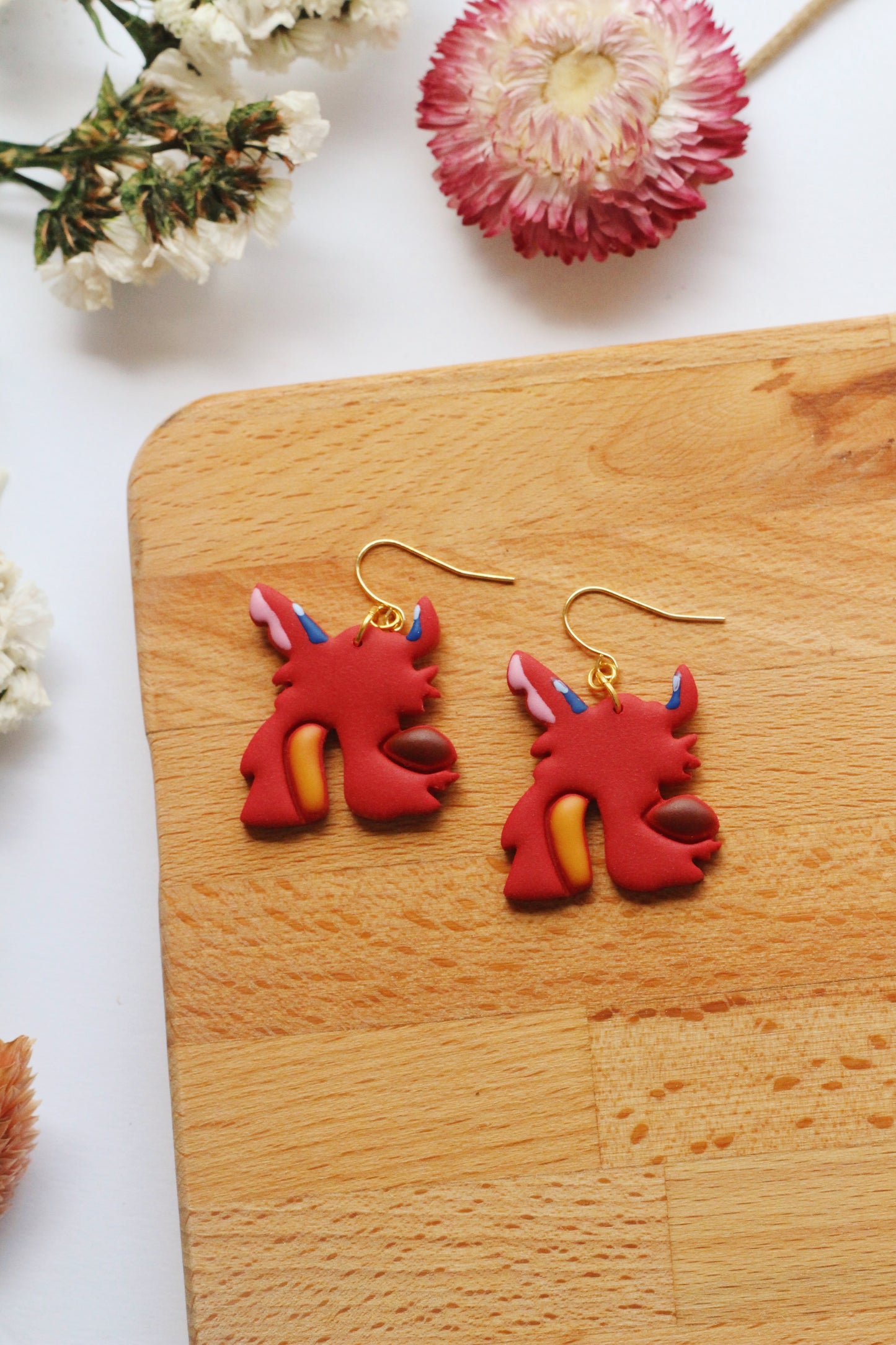 Red dragon-shaped earrings on a wooden board with flowers in the background