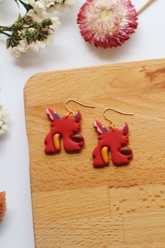 Red dragon-shaped earrings on a wooden board with flowers in the background