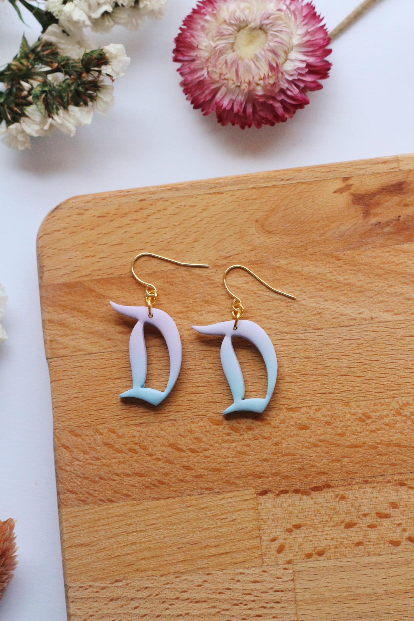 Pair of earrings with 'D' design on a wooden board with flowers in the background